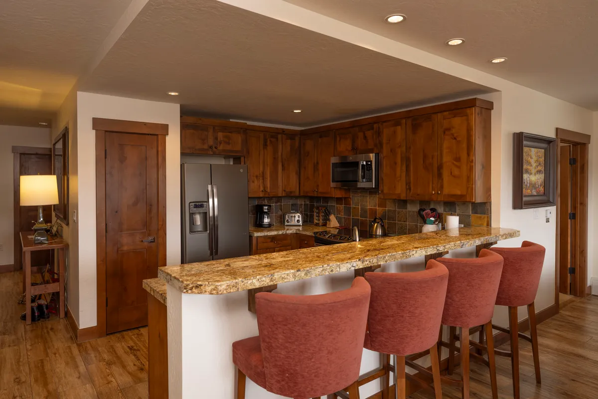 This image centers on a chef’s kitchen with a granite countertop, bar stools for casual seating, warm wood cabinetry, and modern appliances. People are seen socializing and participating in meal prep, evoking a lively and communal atmosphere.
