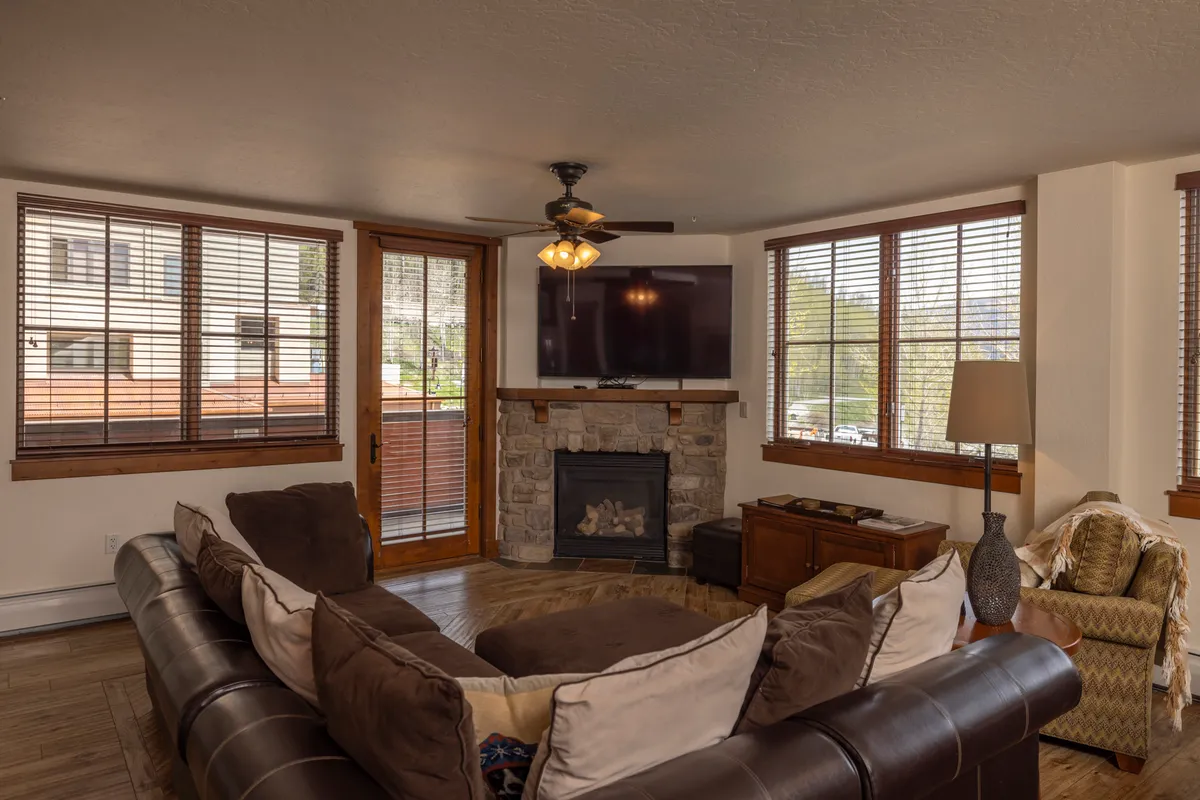 A welcoming living area with a large sectional sofa, a cozy fireplace, and expansive windows with wooden blinds. The television and comfortable seating create an inviting space for gathering, relaxing, or enjoying a family movie night after a day of adventure.