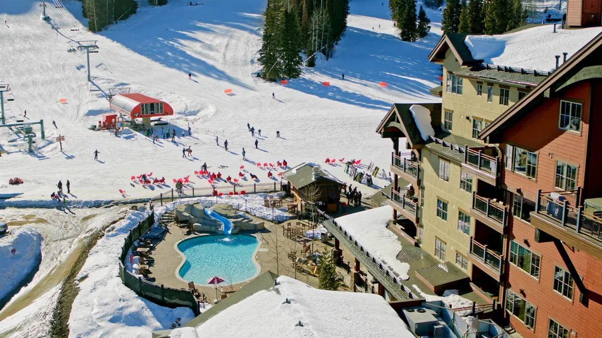 This vibrant exterior scene features snow-covered ski slopes bustling with skiers and snowboarders, a heated swimming pool and hot tub steaming in the foreground, and the welcoming lodge architecture as a backdrop. The lively energy and snowy setting directly convey the slopeside access and après-ski relaxation described in the section.