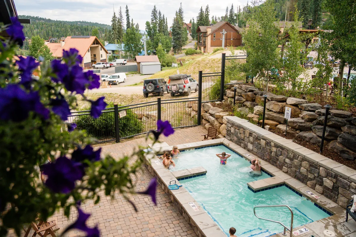 The outdoor heated hot tub is surrounded by snowy landscape and residential lodge buildings, with flowering shrubs peeking through the snow. The space invites post-slope relaxation and socializing in a scenic setting.