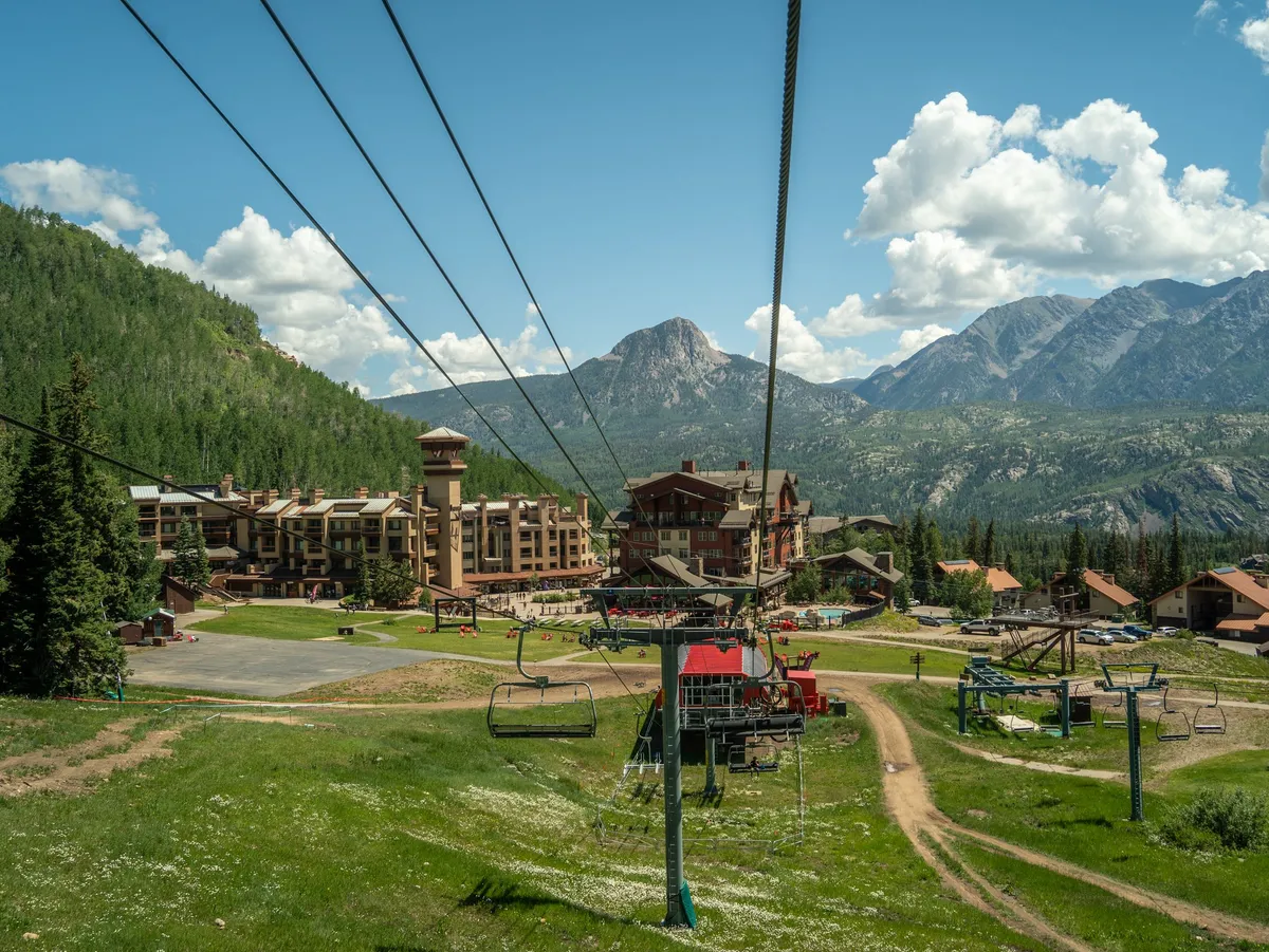 An exterior view of the resort featuring a ski lift set against a lush, green mountain range, with the lodge and open grassy areas visible under a partly cloudy sky. The landscape is vibrant, evoking summer with accessible trails and open space for outdoor activities.