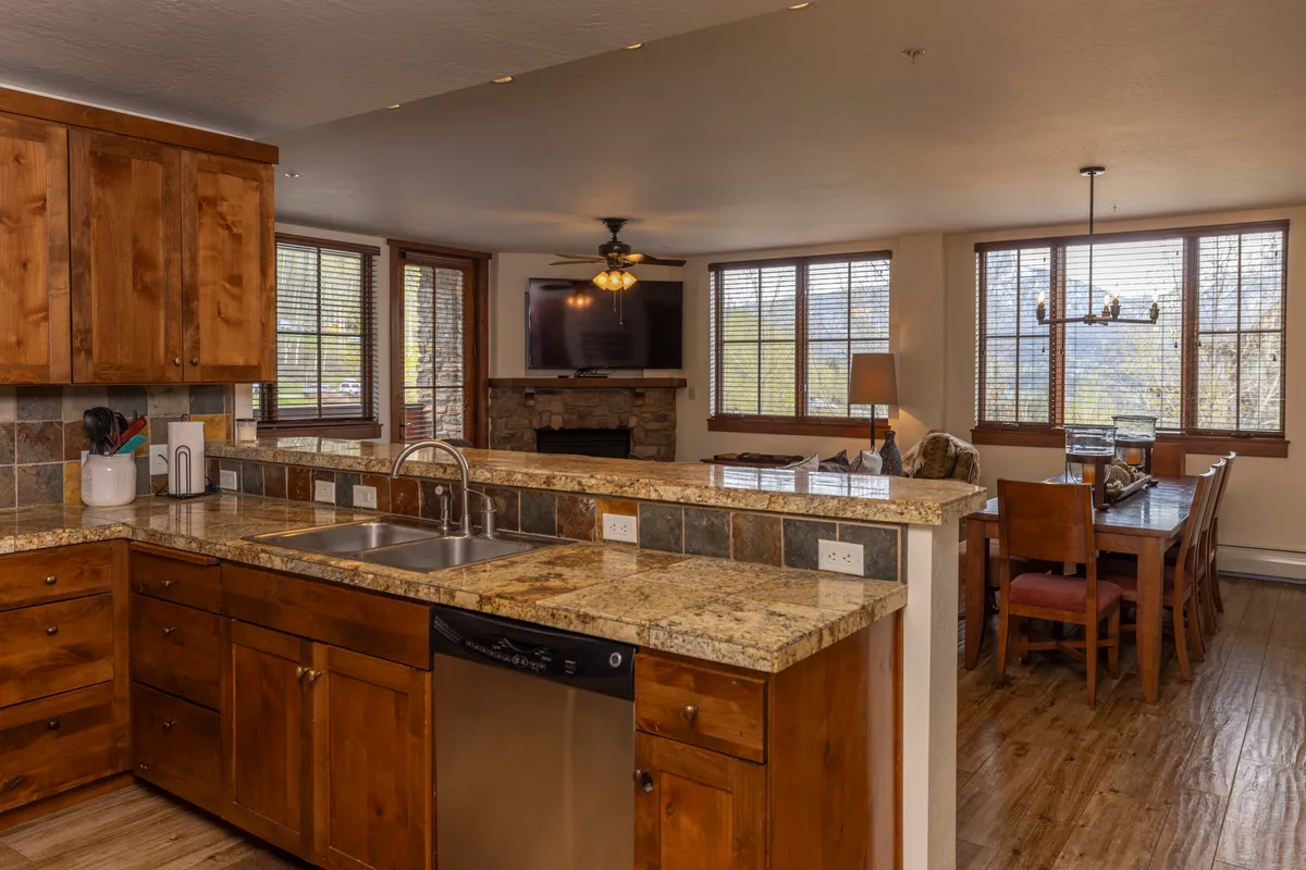 The image showcases a warm, inviting open-concept living space featuring sleek granite countertops, rich wood cabinetry, a spacious dining table, and a cozy fireplace. Sunlight pours in through large windows, framing mountain views and creating the perfect setting for family gatherings or relaxing evenings after a day on the slopes.