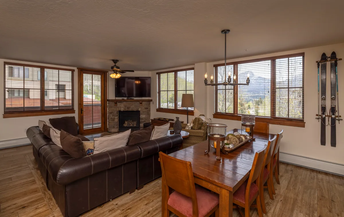 This image captures a warm, open-concept living and dining area featuring a large sectional sofa, a spacious dining table, a cozy fireplace, and expansive windows offering a scenic view of the mountains. The decor includes ski equipment on the wall, emphasizing the lodge’s mountain retreat theme.