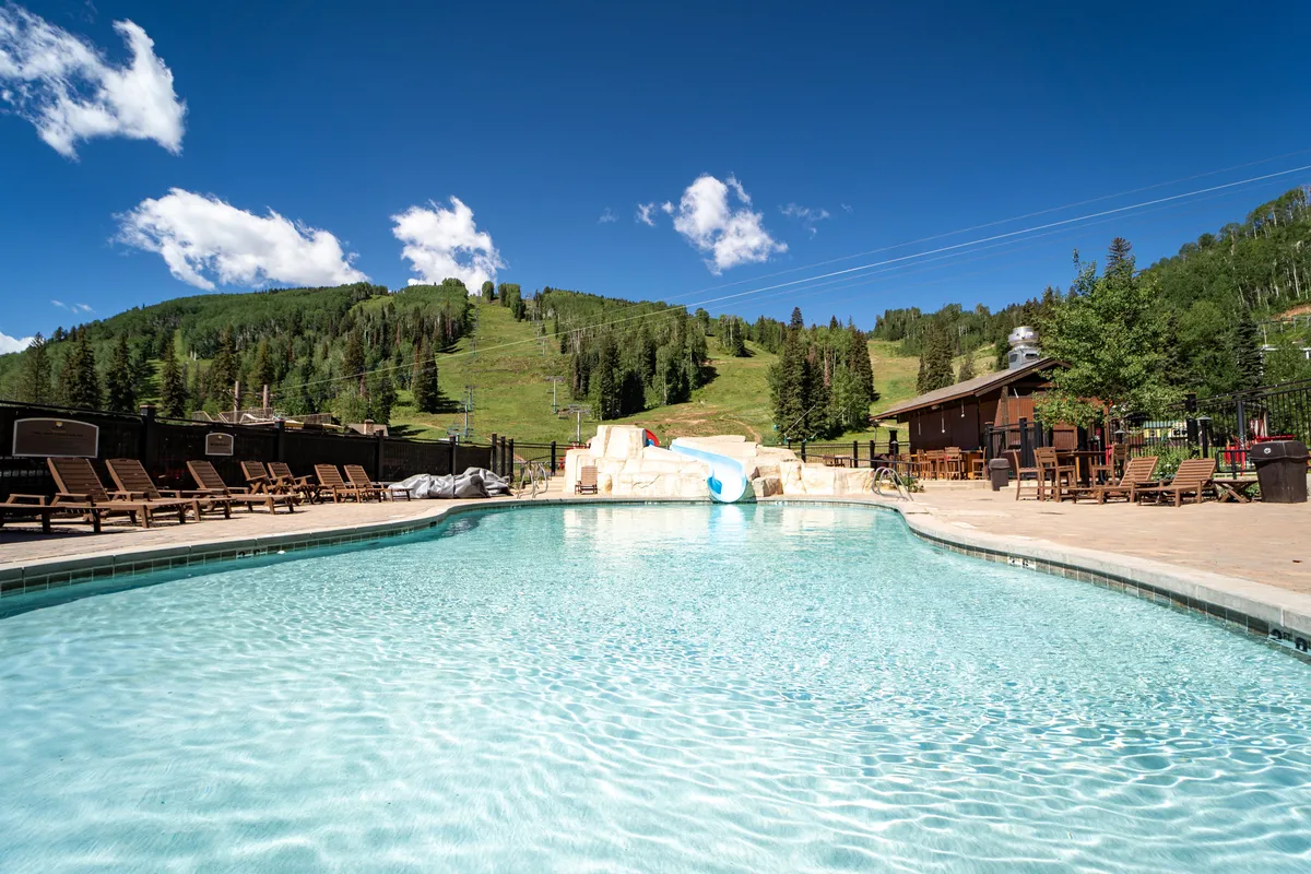 A vibrant outdoor swimming pool framed by lounge chairs and a poolside bar, set against a striking mountain backdrop. The inviting blue water and the pool slide suggest opportunities for both relaxation and fun, with ample space for socializing or enjoying a quiet retreat. The scene is bathed in natural light, enhancing the leisurely and carefree atmosphere.