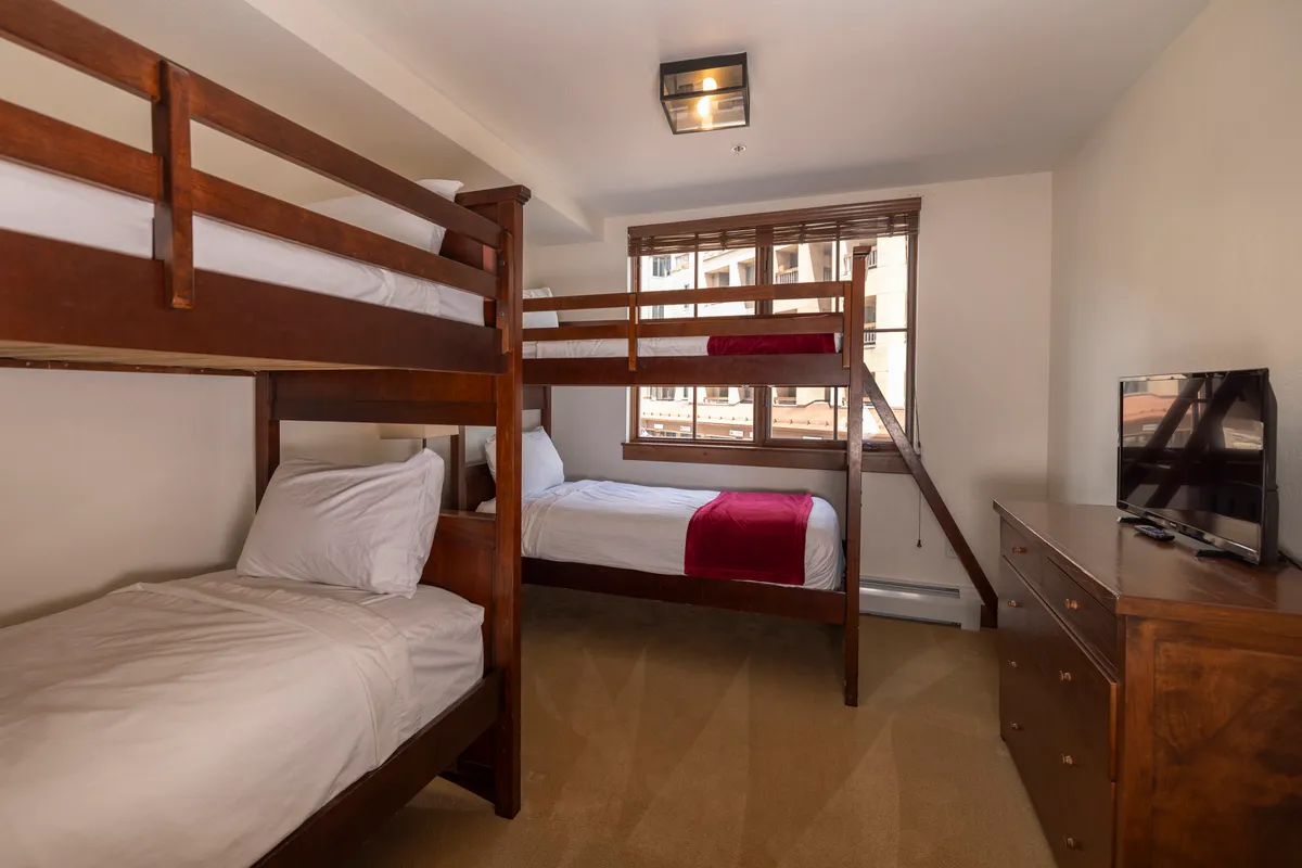 A cozy bunk bedroom designed with twin bunk beds, a single bed, a wooden dresser, a television, and a window letting in natural light. The room is playful yet comfortable, ideal for children or friends, with enough space for relaxing and entertainment.