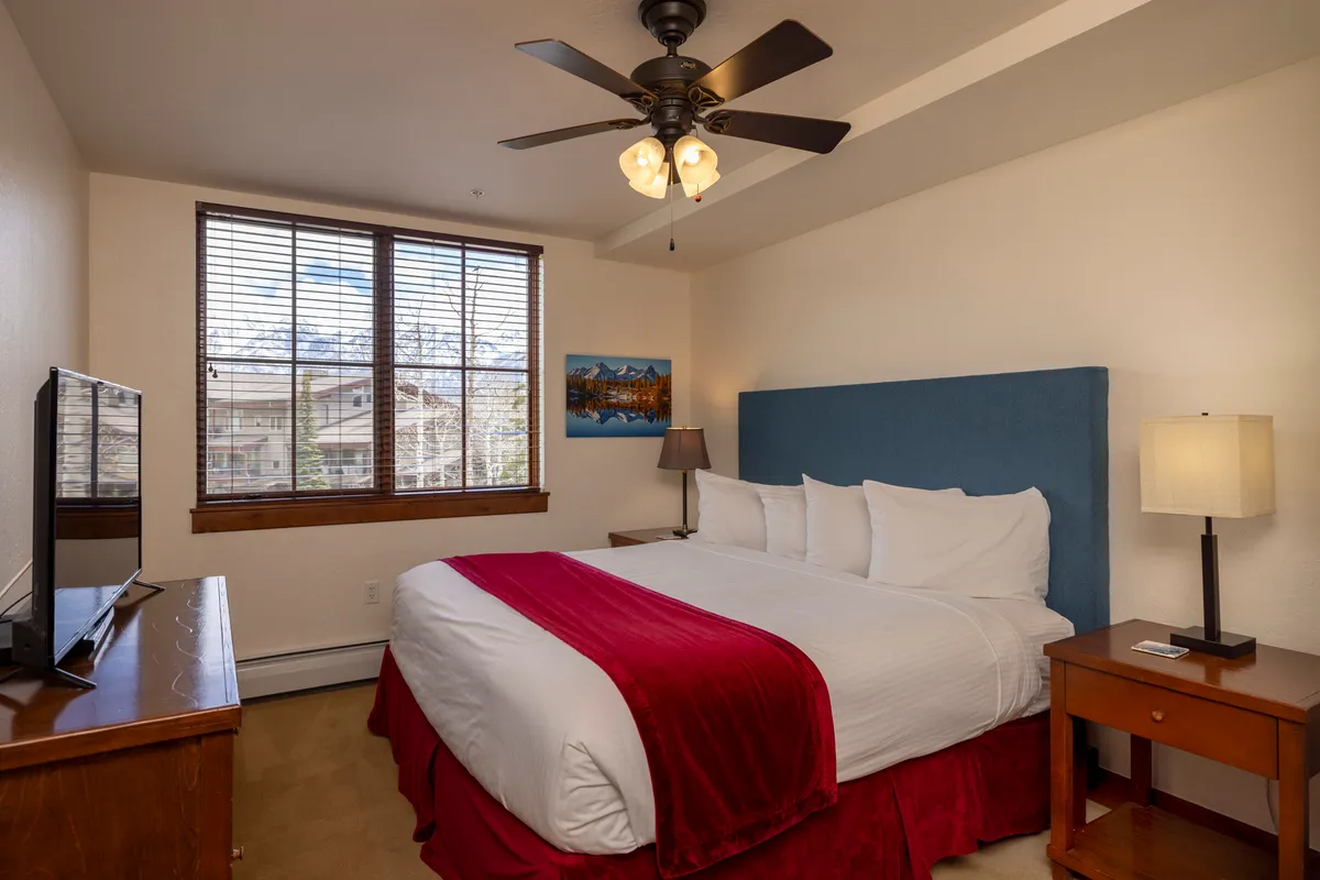A spacious king bedroom featuring a king-sized bed adorned with a red throw blanket, wooden furniture, a large window with blinds, and mounted artwork on the wall. The room exudes a restful and comfortable atmosphere, with abundant natural light and a calming color palette.